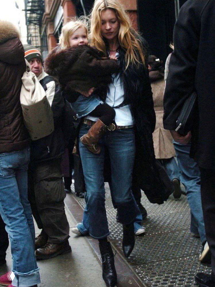 Модель в тёмной шубе держит ребёнка на руках на тротуаре, вокруг много прохожих и городская суета — повседневный уличный момент в холодный сезон.