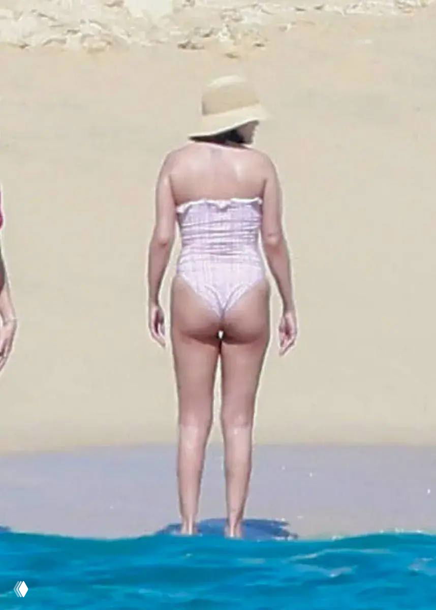 Кадр с пляжа: женщина в розовом купальнике и шляпе показана со спины на песчаном берегу, фотография сделана с расстояния папарацци