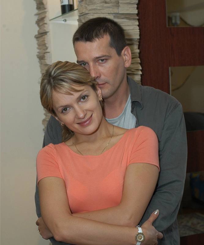Пара обнимается в интерьере: женщина в коралловой блузке в кадре, мужчина стоит позади и нежно держит её за талию.