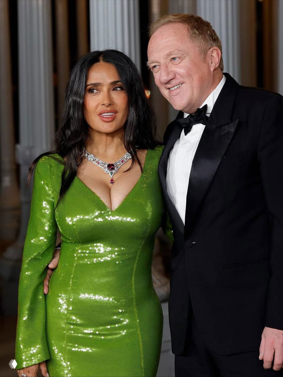 Сальма Хайек рядом с гостем в смокинге на вечере LACMA; зелёное платье блестит и выделяется на фоне архитектурных колонн зала.