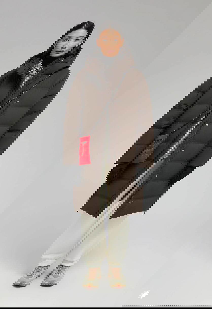 Модель в длинном стёганом коричневом пуховике с капюшоном, белыми спортивными брюками и яркой красной сумкой — комплект для очень холодной погоды.