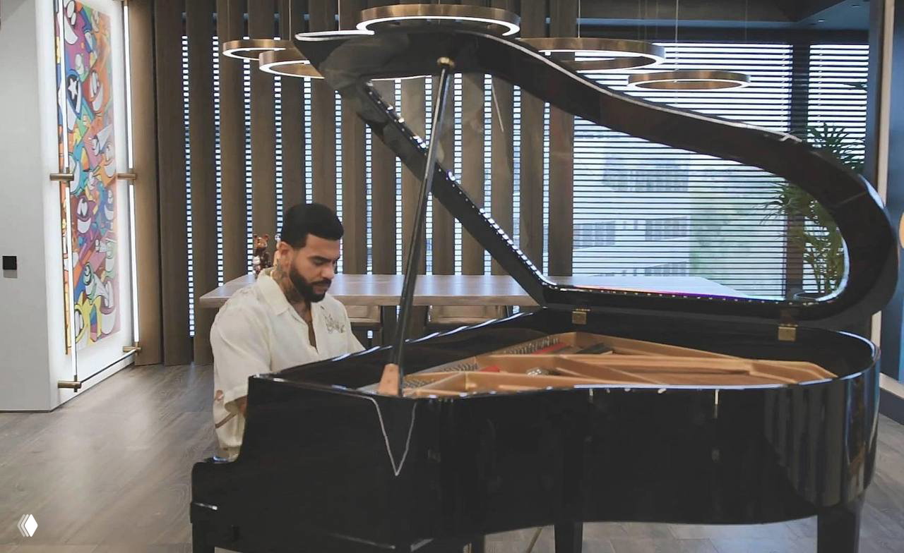 Человек сидит за чёрным роялем в просторной комнате с высокими окнами и декоративным витражом на стене