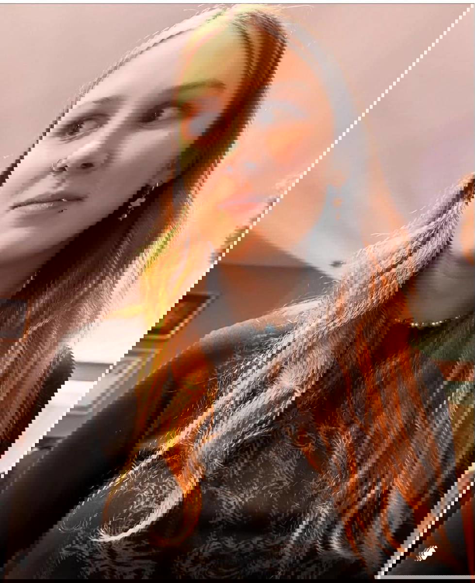 Подростковая девушка в тёмной одежде и с украшениями стоит в помещении; кадр атмосферный, предположительно семейное событие в доме.