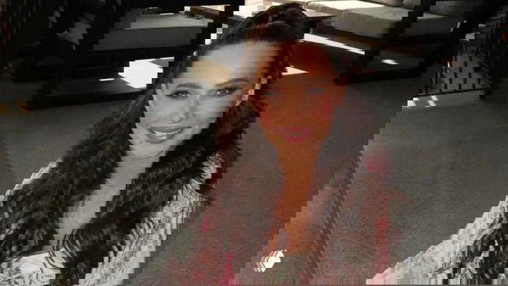 Женщина с тёмными длинными волосами в лёгком платье, сидит в интерьере; мягкое боковое освещение, поза в три четверти и лёгкая улыбка.