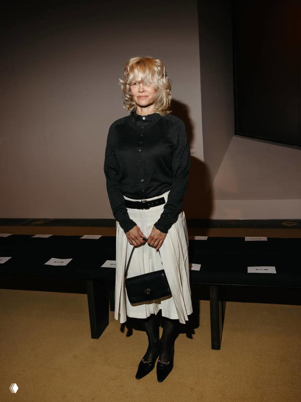 Памела Андерсон сидит на скамейке в зале показа Tory Burch в лёгком макияже и с сумкой в руках, кадр изнутри шоу на Неделе моды.