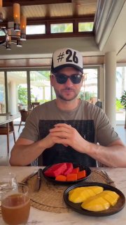 Мужчина в кепке и солнцезащитных очках сидит за столом: на столе тарелки с сезонными фруктами и напиток.