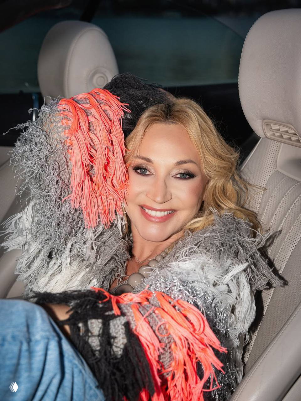 Крупный портрет Кристины Орбайкате в машине: светлый макияж, широкая улыбка и текстурная разноцветная вязаная накидка с бахромой.