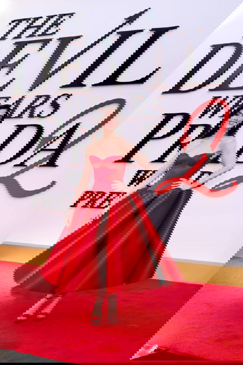 Энн Хэтэуэй в бордовом платье‑корсете и туфлях на высоком каблуке позирует на красной дорожке перед логотипом «The Devil Wears Prada 2».