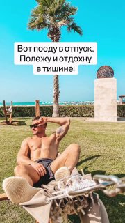 Человек отдыхает на лужайке у моря в шортах; пальмы, яркое небо и строения курорта видны на заднем плане.