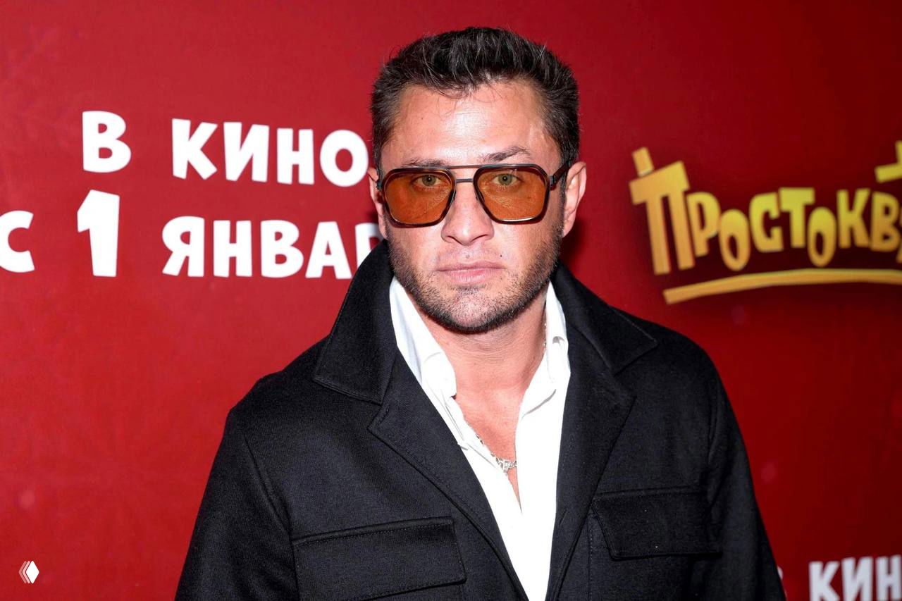 Крупный портретный кадр мужчины в тёмных солнцезащитных очках и пиджаке на красной фотозоне; фон — рекламный баннер кинособытия.