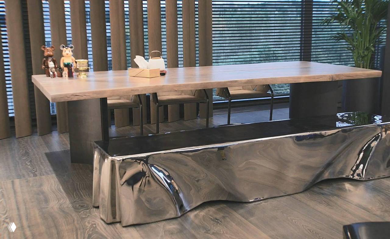 Массивный деревянный стол и блестящая металлическая скамья на фоне больших окон и растений в столовой зоне современной квартиры