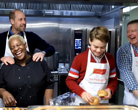 Сцена в кухне: принц Джордж и волонтёры в фартуках готовят и улыбаются, видна посуда и рабочие поверхности центра The Passage в Лондоне.