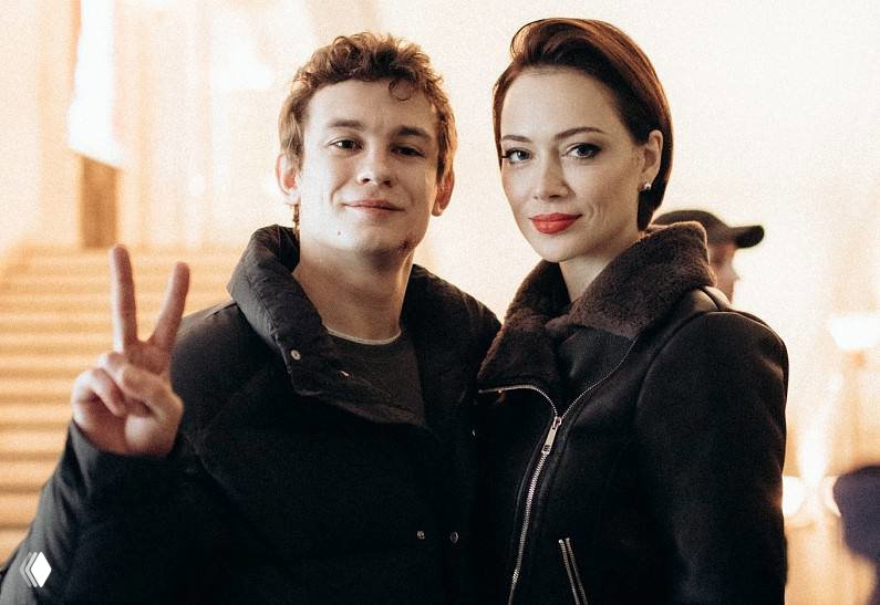 Два человека позируют на лестнице: один показывает жест «V», второй рядом в тёмном пальто; фон интерьер с лестничным маршем.