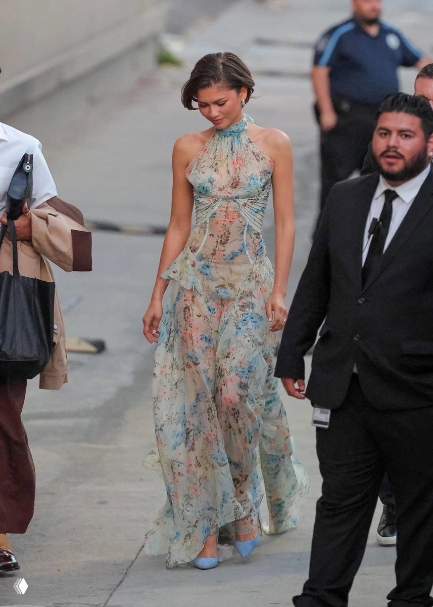 Зендея в полупрозрачном длинном платье с нежным цветочным принтом на съемочной локации промо-тура; уличный кадр в окружении команды, хорошо видна фактура ткани.