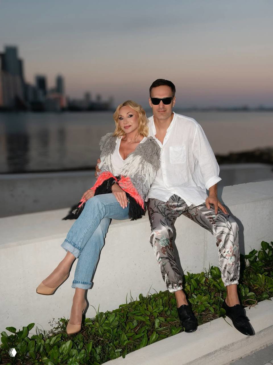 Кристина Орбайкате и мужчина сидят на бетонной ограде у воды в Майами на закате: городской горизонт и спокойная вода на заднем плане.