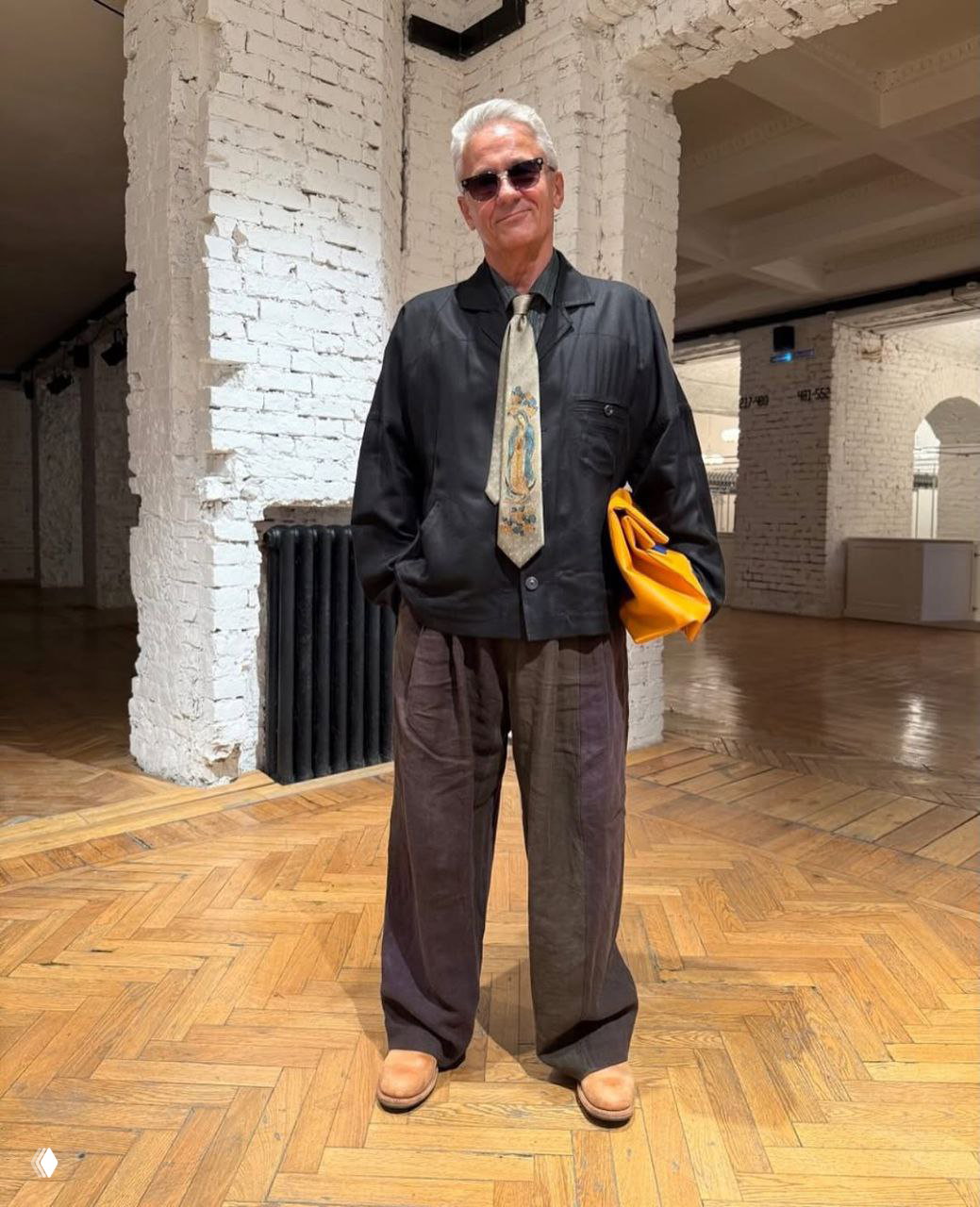 Образ с галстуком: тёмная рубашка и свободные тёмные брюки, жёлтая сумка‑конверт и солнцезащитные очки в интерьере с белыми колоннами.