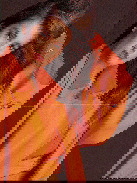 Ретро‑портрет модели в оранжевой рубашке в расслабленной позе, кадр подчёркивает макияж, цветовую палитру и гламурную эстетику 1990‑х.