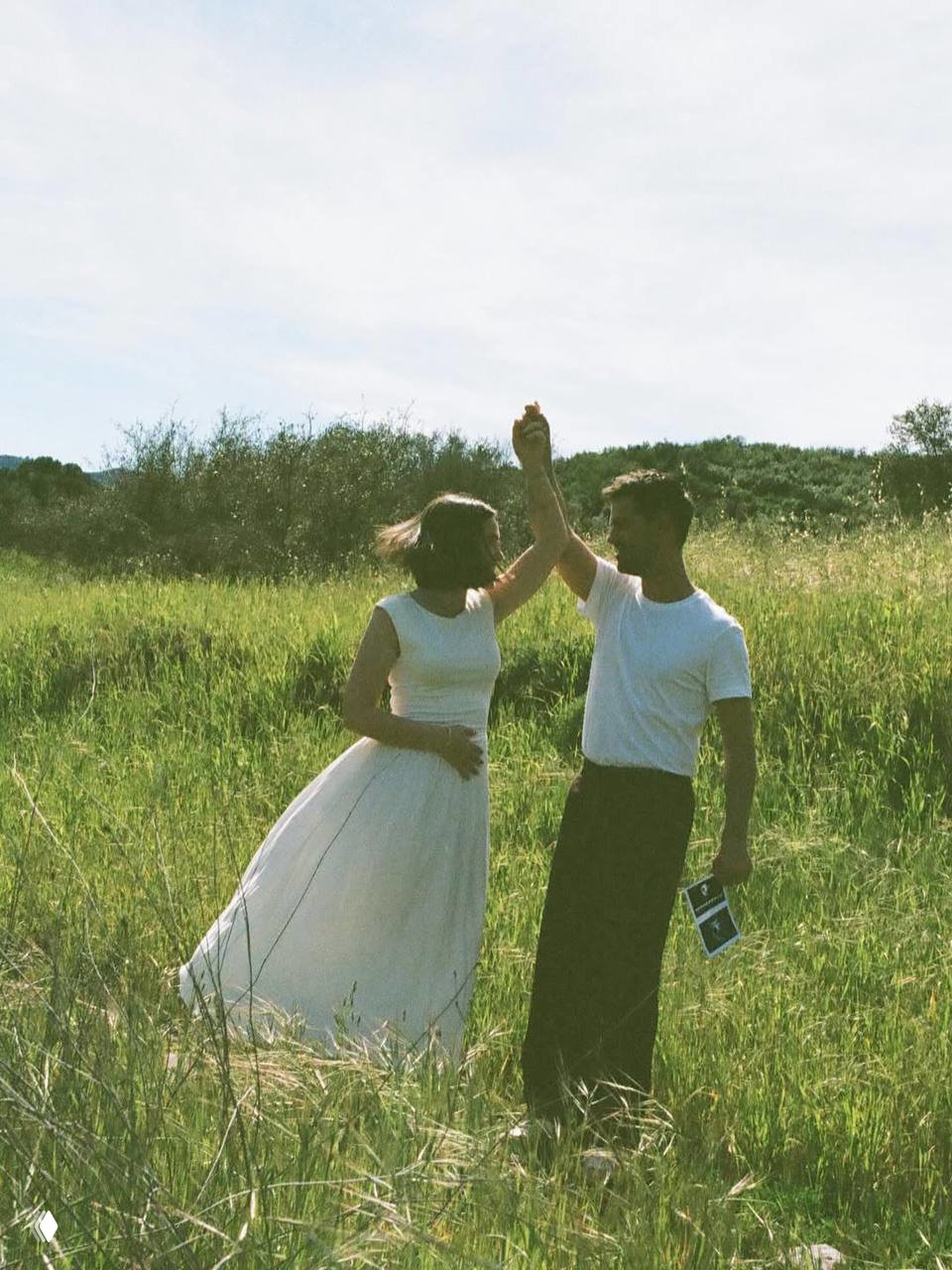 Та же пара танцует на траве: мужчина держит руку женщины, она в лёгком платье, он держит снимки — кадр беременности на природе.