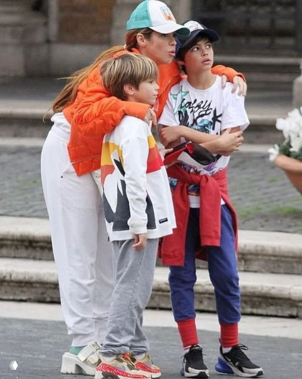 Уличный семейный кадр: женщина в кепке обнимает двоих детей, они в повседневной одежде, прогулка по городу и внимание друг к другу.