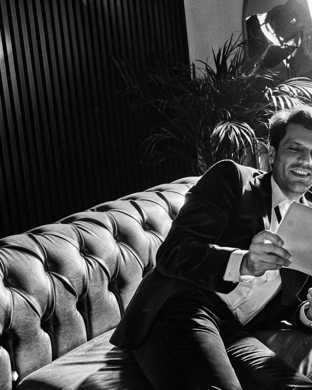 Каан Урганджиоглу в чёрном костюме улыбается и листает бумаги, сидя на дублёном кожаном диване в студии — кадр в чёрно‑белой гамме.