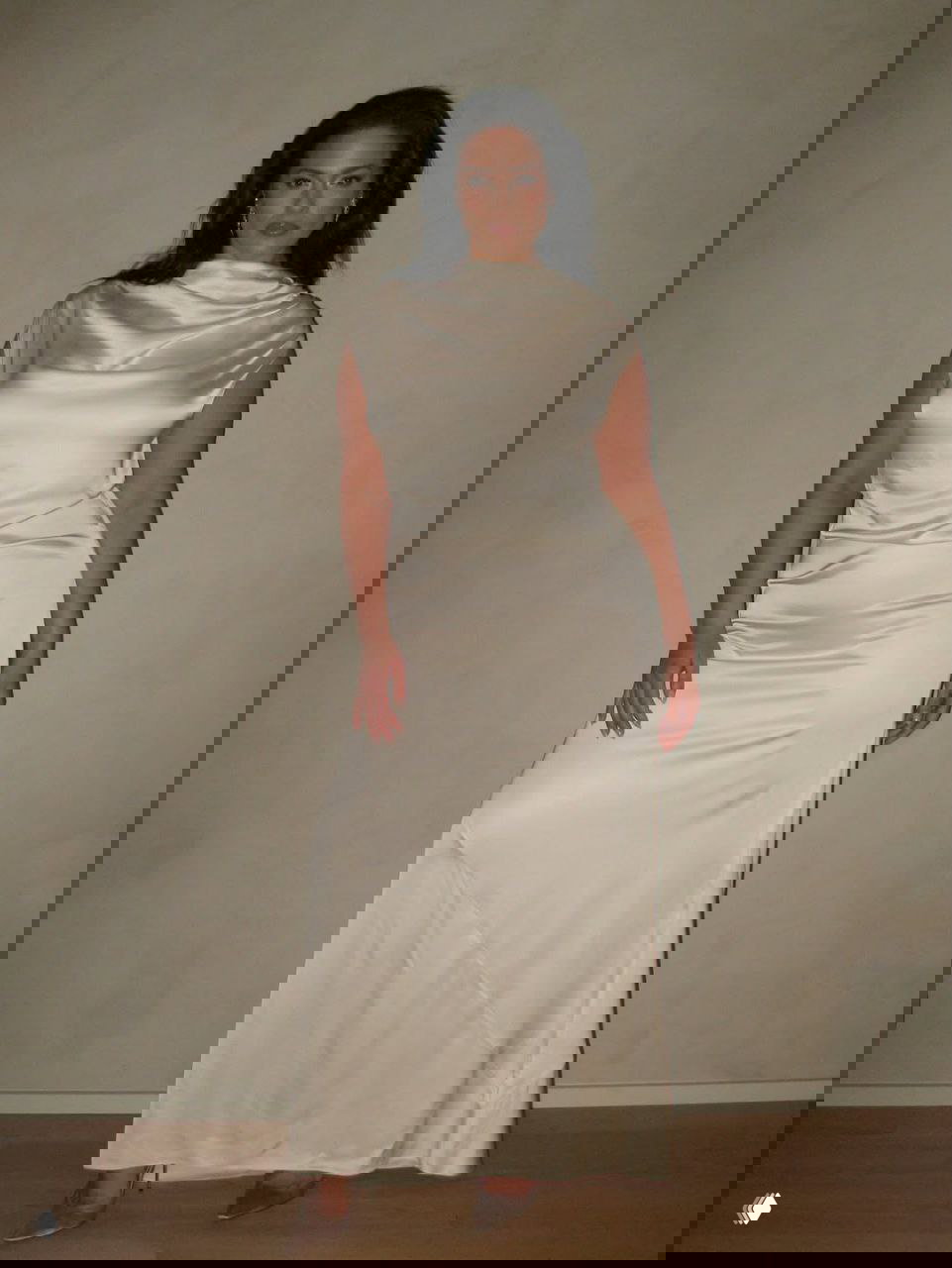 Женщина в длинном молочно‑атласном платье в полный рост на фоне нейтральной стены, демонстрация силуэта и посадки платья.