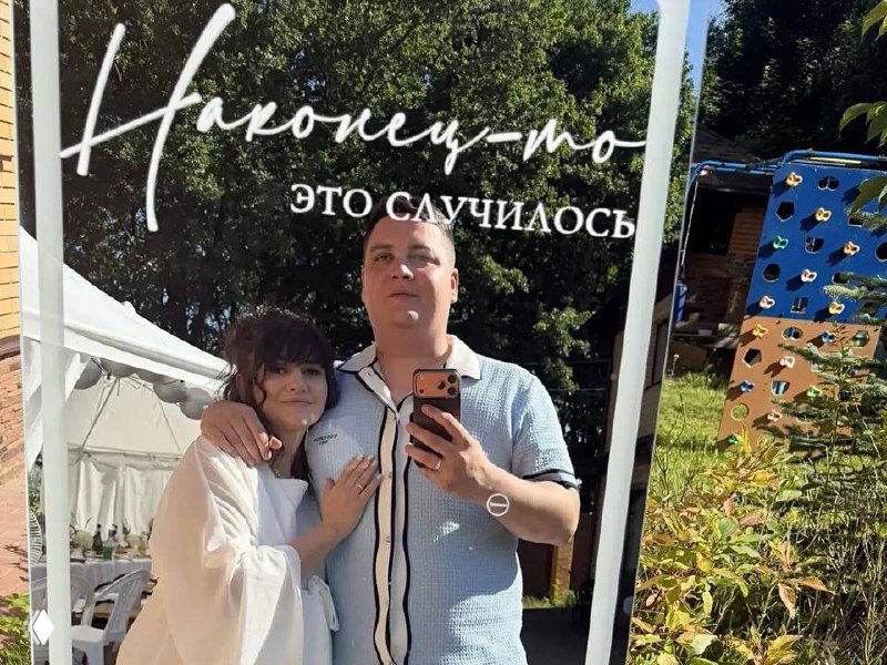 Анна Руднева и муж делают селфи у зеркальной фотозоны с надписью «наконец-то — это случилось» на фоне летнего сада.