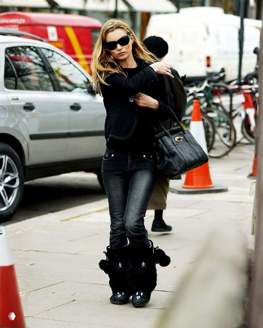 Фигура в чёрном пиджаке и тёмных джинсах с низкой талией, большие сумки и солнцезащитные очки на фоне городской улицы — стритстайл в движении.