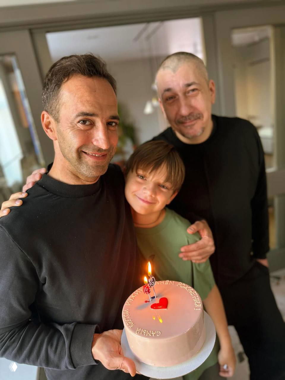 Группа: мужчина держит торт, рядом ребёнок и ещё один взрослый, все улыбаются — семейный тёплый кадр с праздничной свечой на торте.