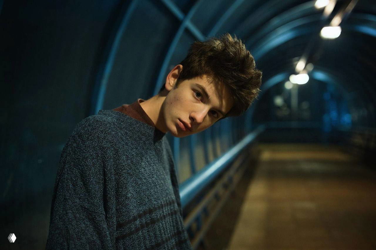 Молодой мужчина в тёмном свитере на пешеходном мосту или тоннеле, атмосферный вечерний кадр с боковым освещением и размытой перспективой за спиной