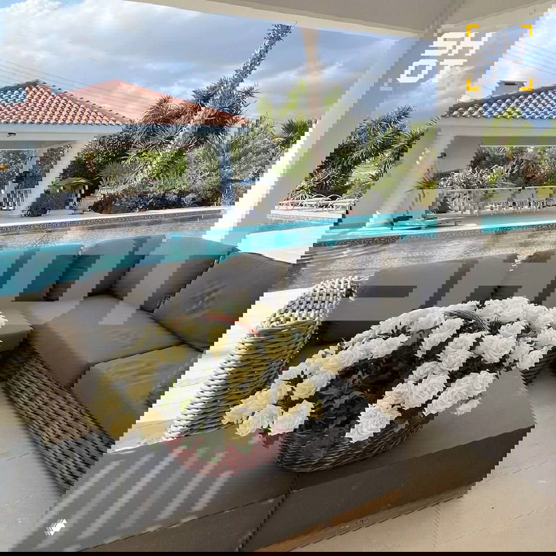 Терраса у бассейна с плетёной уличной мебелью и большой корзиной белых цветов на столике; видна часть бассейна и летняя кухня на заднем плане