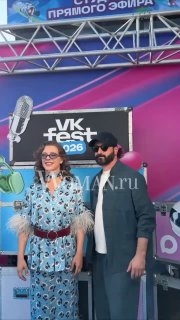 Двое людей на синей дорожке VK Fest 2026 — женщина в платье с принтом и мужчина в тёмной одежде на фоне сцены.