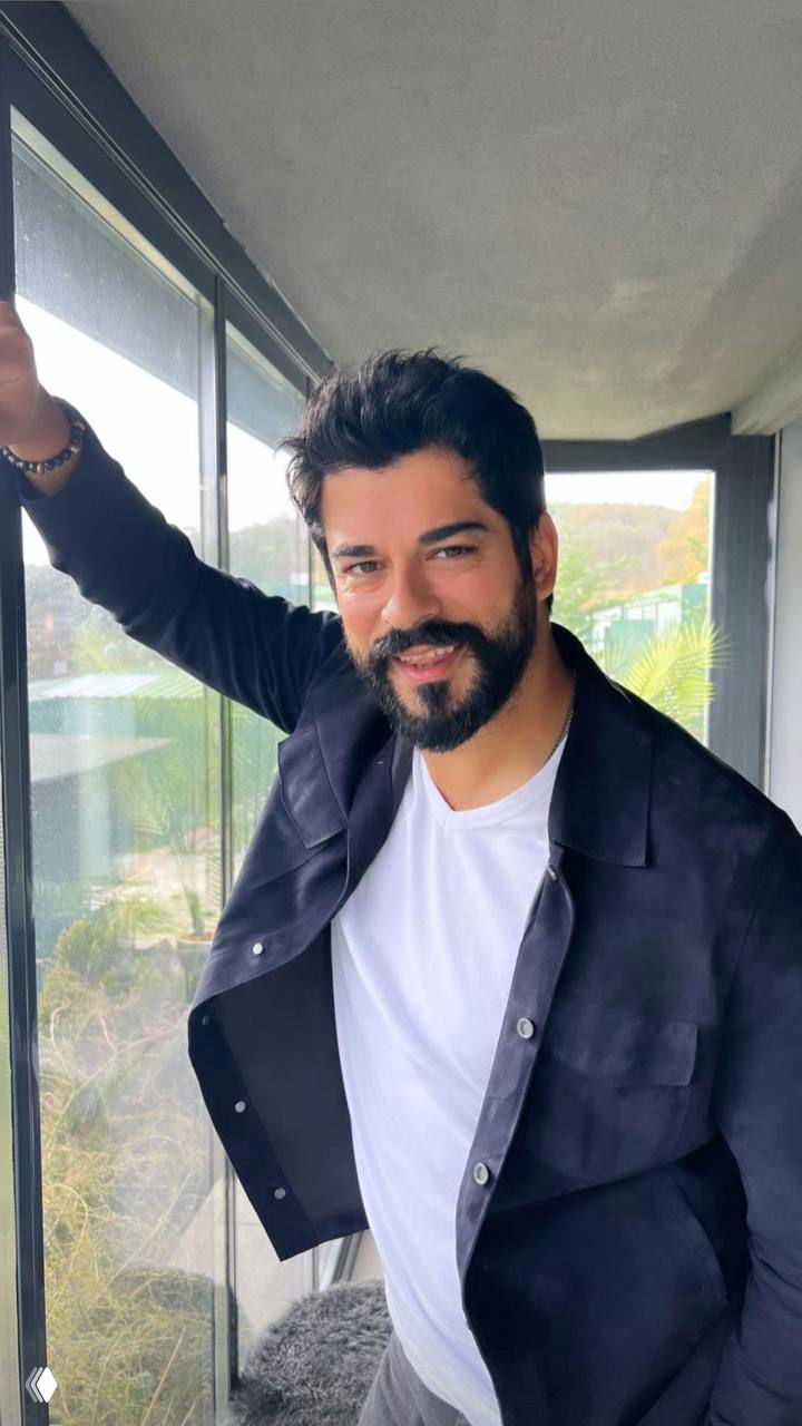 Мужчина в повседневной одежде стоит у окна-балкона, лёгкая улыбка, неформальный портрет на фоне зелёных холмов и городской террасы.