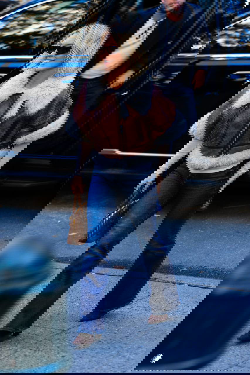 Актриса в короткой коричневой дублёнке с меховой отделкой и джинсах, сумка в руке и непринуждённая улыбка при выходе из автомобиля