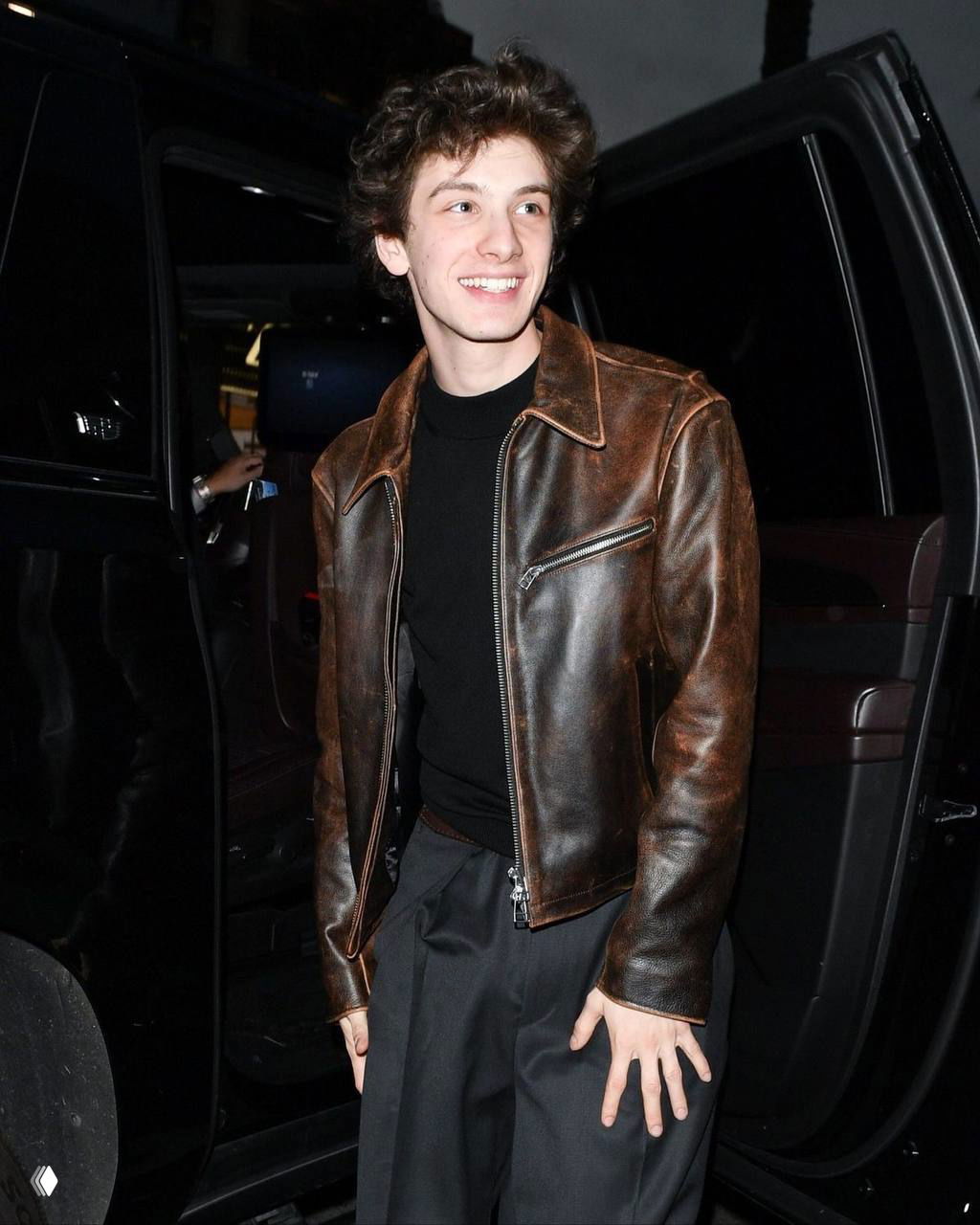 Молодой человек в коричневой кожаной куртке выходит из автомобиля ночью, тёплое освещение, улыбка и непринуждённый стиль, папарацци‑кадр