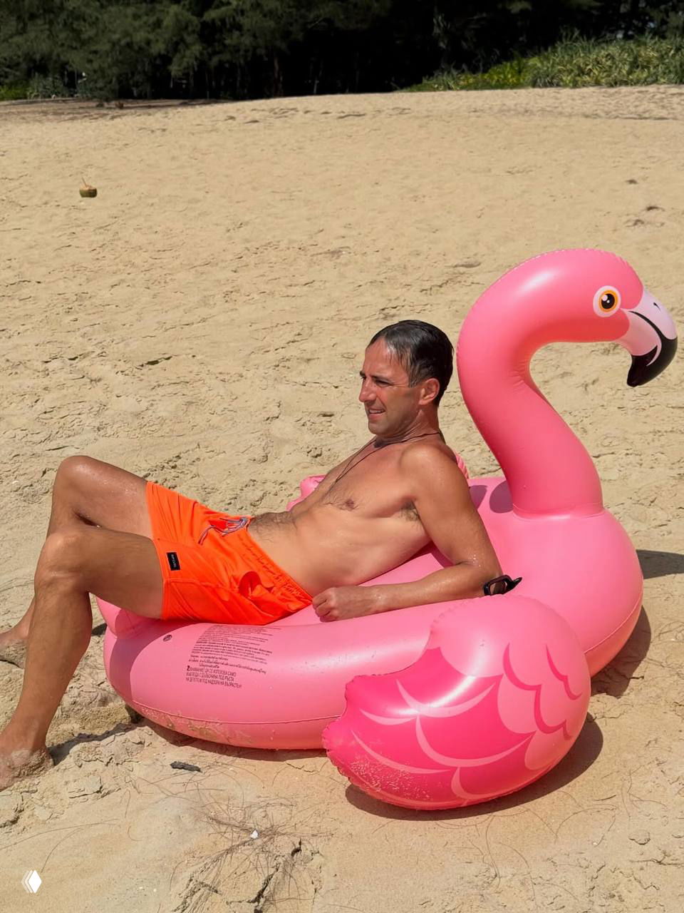 Мужчина отдыхает на пляже, лёжа в розовом надувном круге в форме фламинго; яркий летний кадр и пляжная сцена.