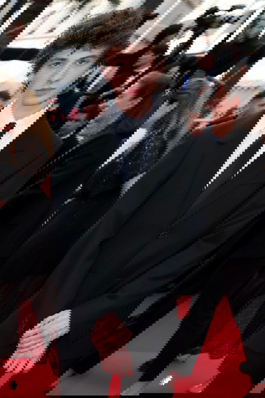 Мужчина в формальной чёрной одежде и галстуке на красной дорожке кинофестиваля, вокруг фотографы и гости, официальный внешний вид и строгая поза