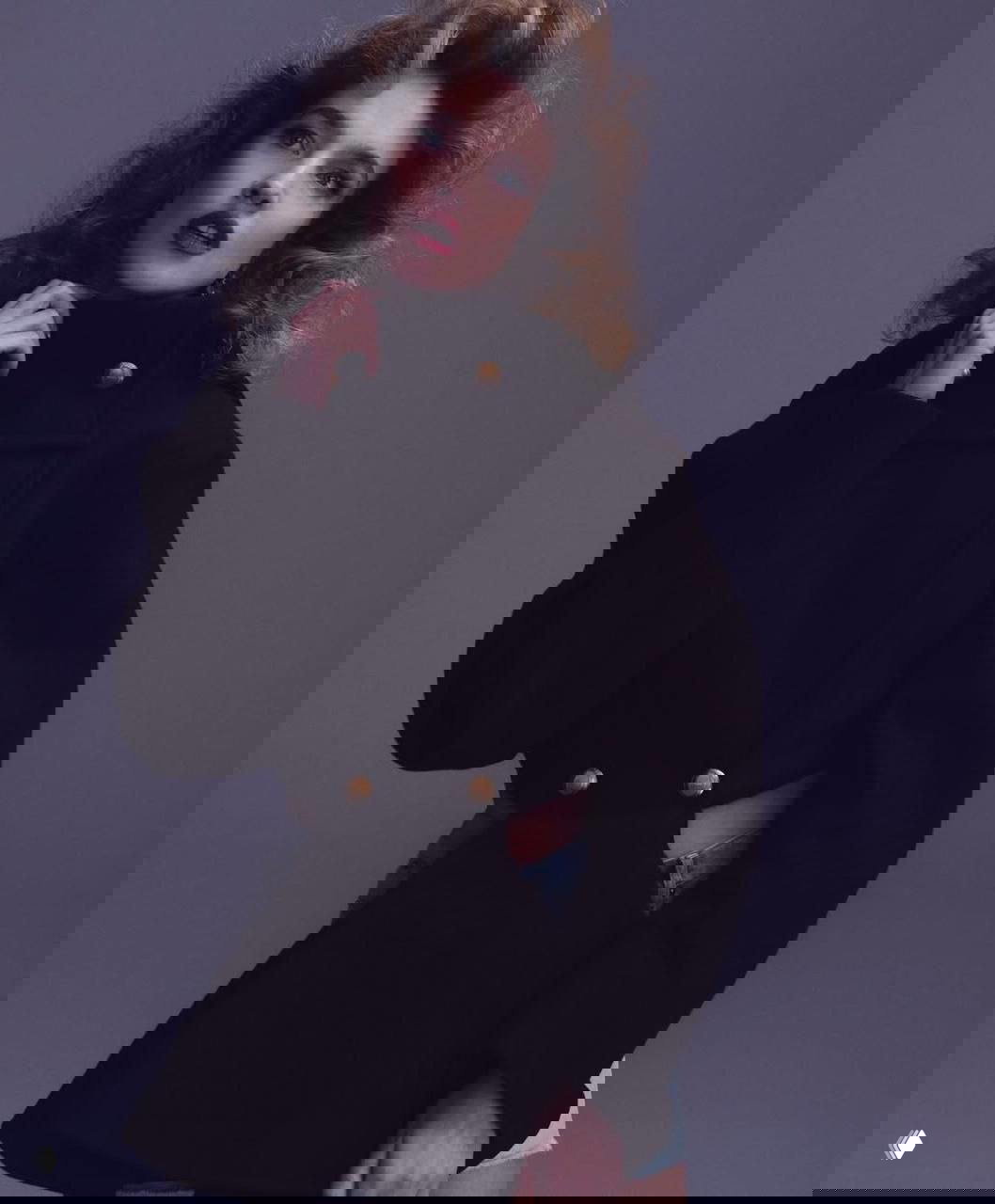 Портретный снимок: женщина в тёмном пальто в студийной съёмке, выразительный макияж и волнистая причёска на однотонном фоне.