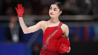 Аделия Петросян в красном костюме на льду, жест рукой и сосредоточенное выражение лица во время выступления на соревнованиях