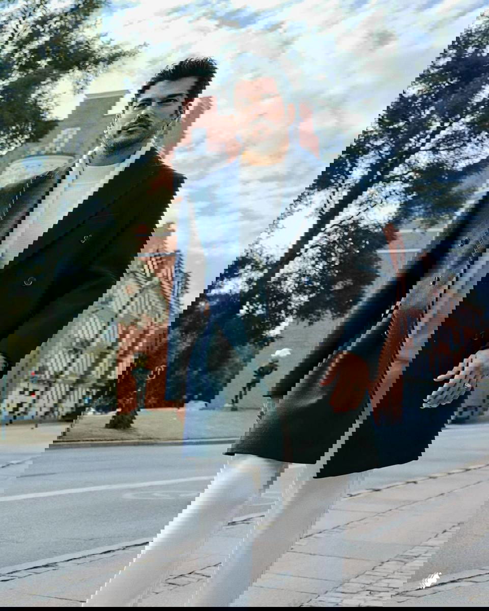 Мужчина в бежевом пиджаке облокотился о перила на фоне городского пейзажа и зелёных растений, спокойная поза и утончённый стиль.