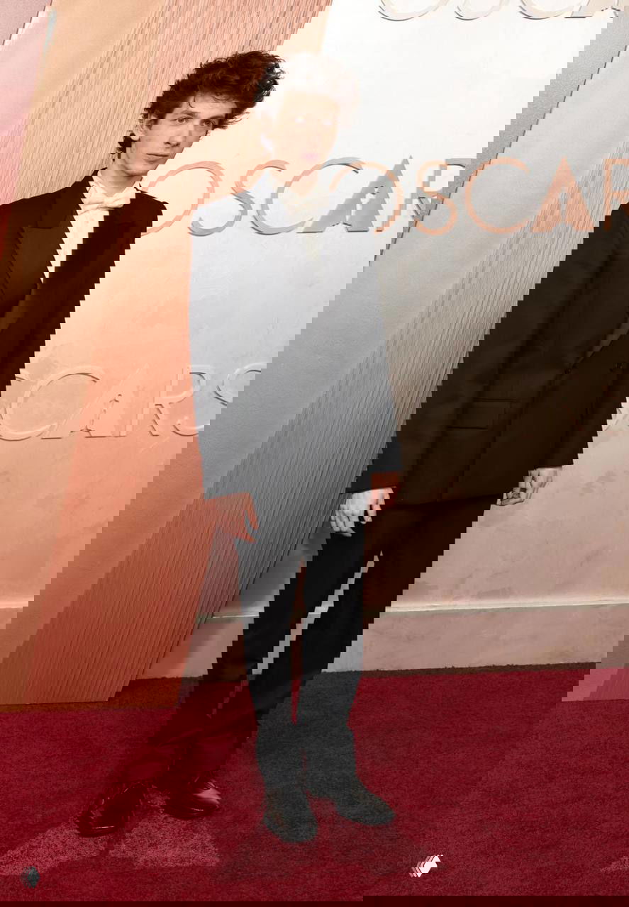 Полный рост молодой мужчины в чёрном смокинге на красной дорожке у фотозоны с логотипами, официальный образ для светского мероприятия и статичная поза