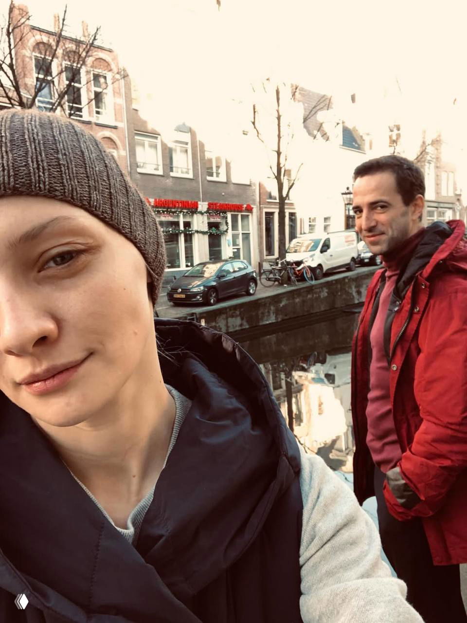 Уличное селфи пары на канале европейского города: женщина на переднем плане, мужчина на заднем плане, городской пейзаж и отражение в воде.