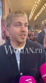 Вертикальный кадр: Сергей Романович общается с прессой, впереди видны микрофоны с логотипом Woman.ru, фон заполнен людьми.