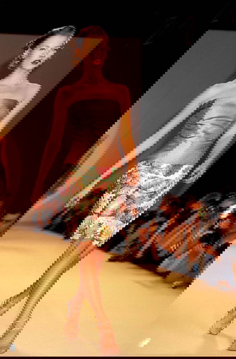 Молодая Кейт Мосс дефилирует по подиуму в романтическом образе с юбкой в цветочный принт, публика и фотографы на заднем плане, классический показ.