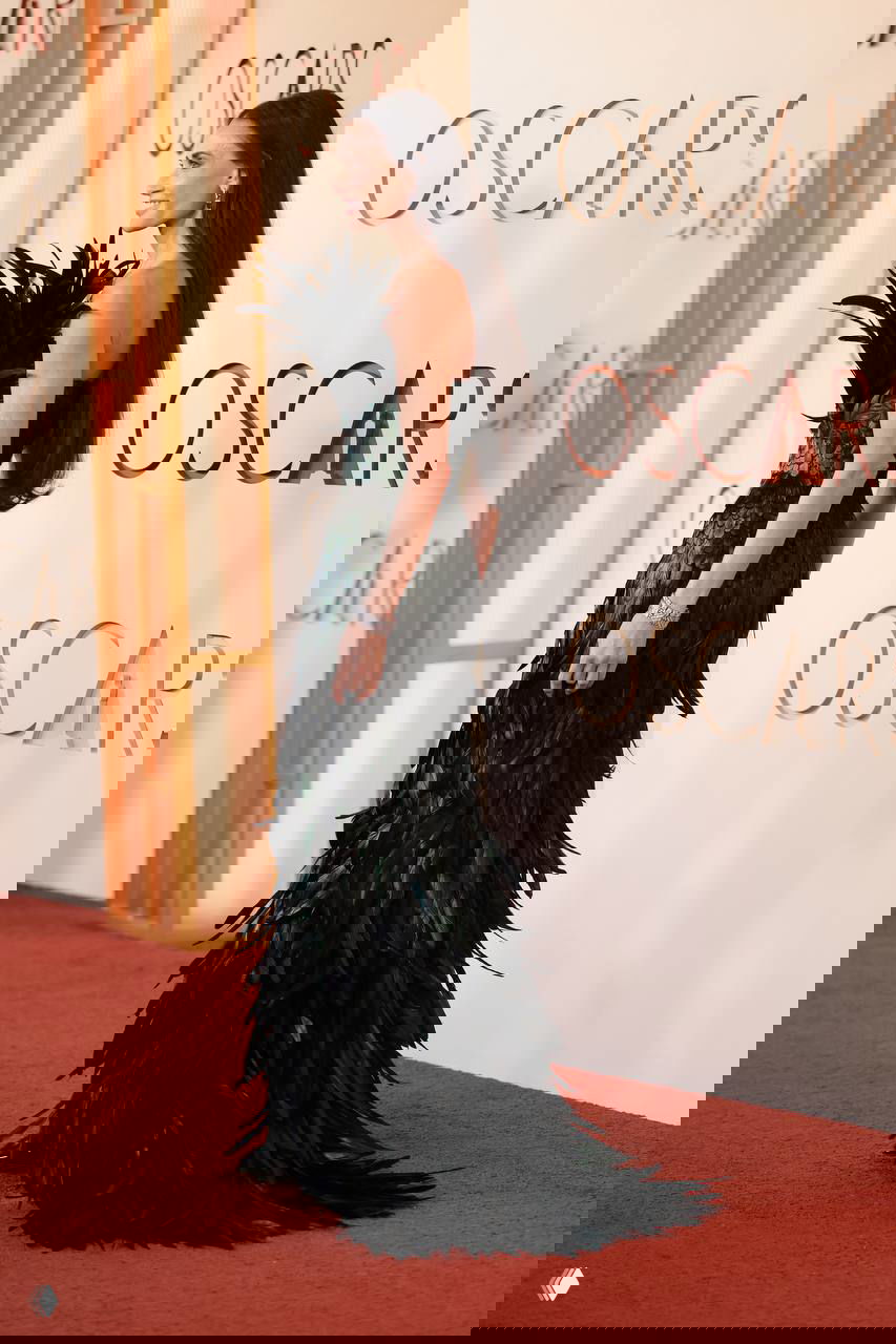 Женщина в длинном чёрном платье с перьями идёт по красной дорожке у пресс-волла OSCARS; виден полный рост и силуэт наряда.