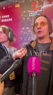 На премьере мужчина в очках даёт интервью журналистам: микрофон Woman.ru на переднем плане, на фоне афиша и люди на красной дорожке