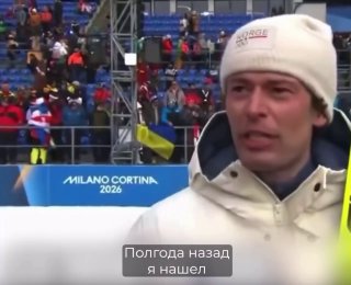 Биатлонист в зимней одежде даёт интервью на фоне трибун и флага; кадр с церемонии Милано-Кортина 2026, награждение