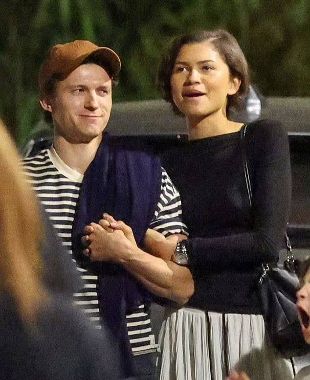 Пара на вечерней прогулке у парковки: мужчина в полосатой футболке, женщина в чёрном топе и длинной юбке, люди вокруг.