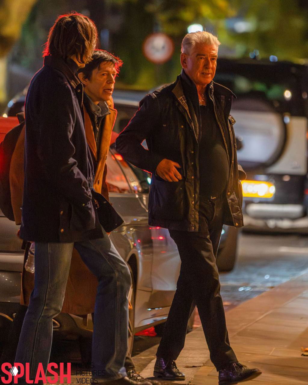 Пирс Броснан и ещё один мужчина идут по тротуару в Лондоне ночью: семейная прогулка после ужина, заметна городская улица и припаркованные машины.