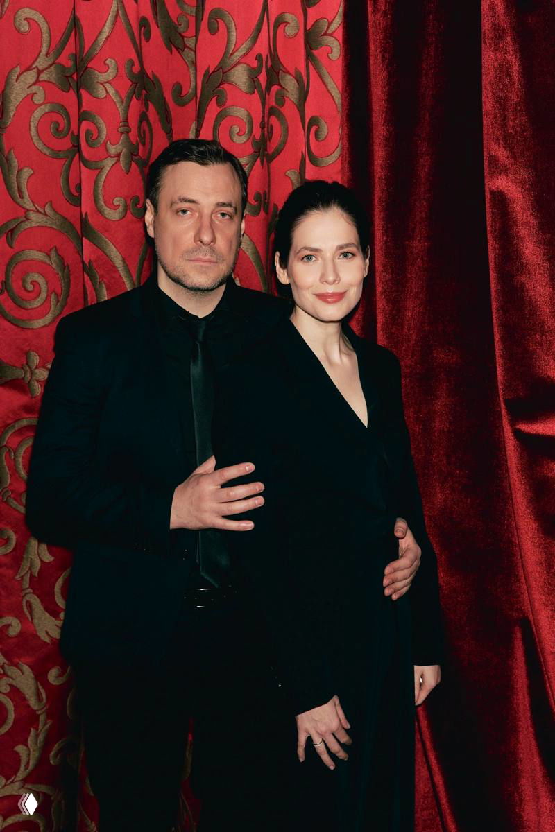 Два человека в тёмных вечерних нарядах позируют на фоне красных штор — снимок из ряда официальных портретов гостей благотворительного вечера.