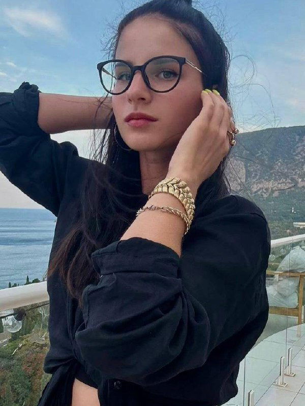 Крупный план молодой девушки в очках на фоне моря и скал; серьёзный взгляд и повседневный стиль в кадре с видом на побережье.